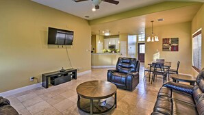 A spacious living room with a ceiling fan, a TV mounted on the wall, a round coffee table, leather sofas, and a kitchen area in the background. - Walk to Ocean: Lakefront Dickinson Stucco House! (Dickinson)