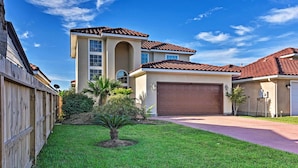 A two-story house with a brown garage door, a small front yard with a palm tree, and a wooden fence. - Walk to Ocean: Lakefront Dickinson Stucco House! (Dickinson)