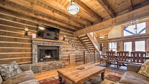 A log cabin interior with a stone fireplace, wooden furniture, and a staircase leading to an upper level. - Hot Tub + Pool Table: Cozy Franklin Cabin! (Franklin)