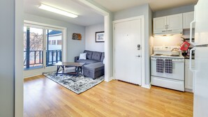A modern kitchen with white cabinets, a stove, and a refrigerator. There is a gray sofa and a coffee table in the living area. - Cozy Water-view Apt in the Heart of Downtown! (Hallowell)