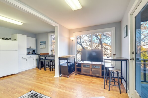A modern kitchen with white cabinets, a dining area with a wooden table and chairs, and a large window with a view of trees.