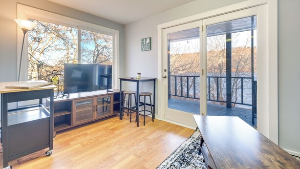 A modern living room with a wooden floor, a TV, a small table with stools, and a large sliding glass door leading to a deck. - Cozy Water-view Apt in the Heart of Downtown! (Hallowell)