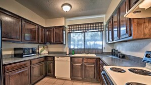 Une cuisine avec des meubles de rangement en bois foncé, un lave-vaisselle blanc, un four à micro-ondes, une cuisinière et une fenêtre agrémentée d’une lambrequin à carreaux.