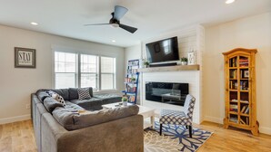 A modern living room with a gray sectional sofa, a white coffee table, a patterned armchair, a bookshelf, a TV mounted above a fireplace, and a wooden bookcase. - Spacious Townhome ~ 7 Mi to Deer Valley Resort! (Heber City)