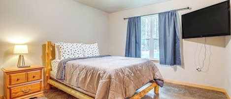 Ein Schlafzimmer mit Bett, Nachttisch aus Holz, Lampe, Deckenventilator, Fenster mit Vorhängen und einem an der Wand montierten Fernseher.