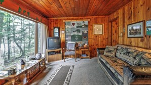Ein gemütliches Blockhaus-Interieur mit Holzwänden, einem großen Fenster mit Blick auf einen Wald, einem Fernseher, einer Couch und einem kleinen Tisch mit Teppich.
