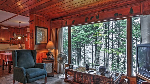 Ein gemütliches Blockhäuschen-Interieur mit Holzbalkendecke, einem großen Fenster mit Blick auf den Wald, einem bequemen Sessel, einem Holztisch mit Ventilator und einem Fernseher.