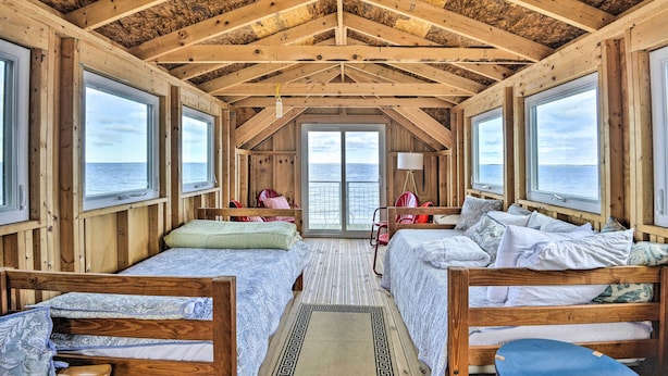 Eine Hütte mit zwei Holzbetten, einem kleinen Tisch und Blick auf das Meer durch große Fenster.