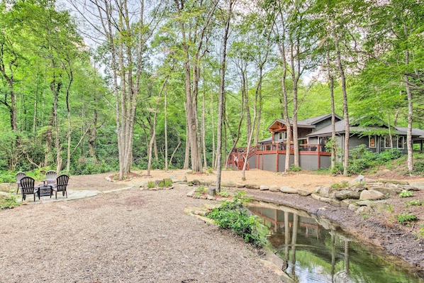 A cabin surrounded by a forest with a small pond, outdoor seating, and a stone pathway.