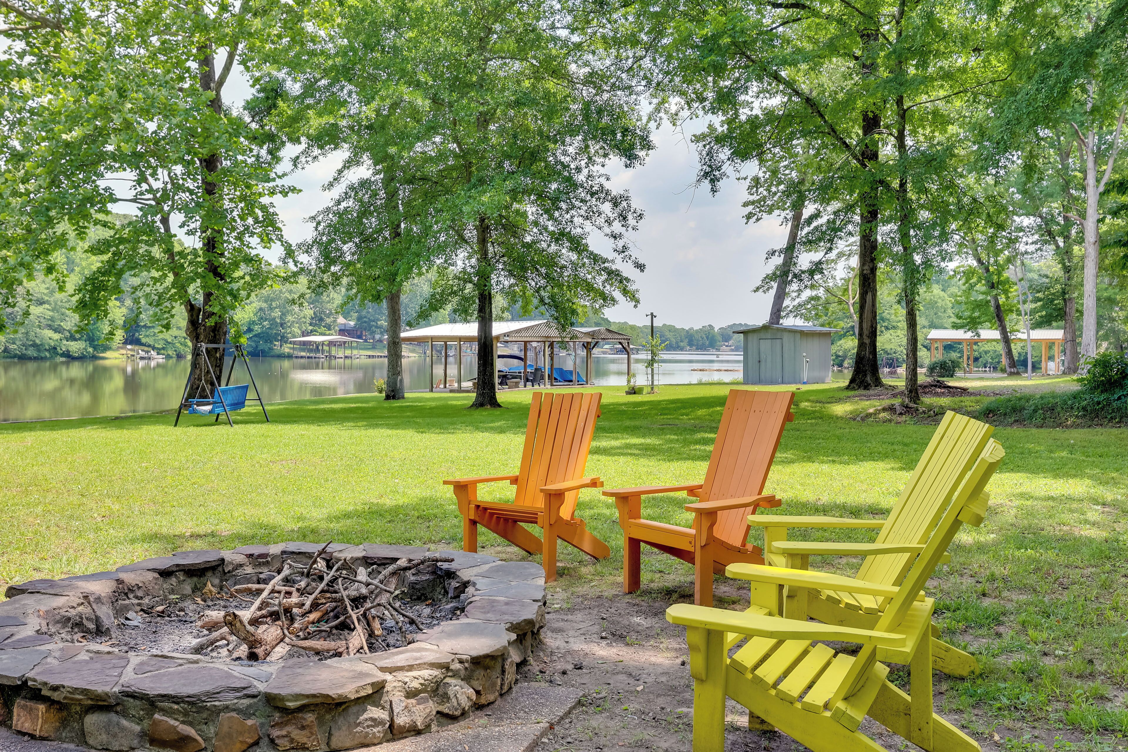 A lakeside outdoor seating area with Adirondack chairs, a stone fire pit, and a grassy lawn.