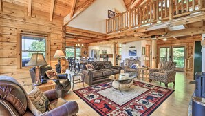 A spacious log cabin interior with a loft bedroom, leather furniture, a large rug with animal prints, and a wood-burning stove. - River Access & Game Room: Piney Creek Cabin! (Piney Creek)