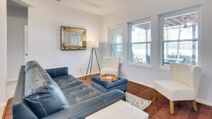 A living room with a dark blue leather sofa, a white armchair, and a small ottoman. There is a floor lamp, a mirror with a gold frame, and large windows with white shutters.