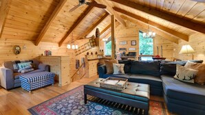 A cozy log cabin interior with a wooden ceiling, a staircase, a living room with a sofa and ottoman, and a kitchen area.