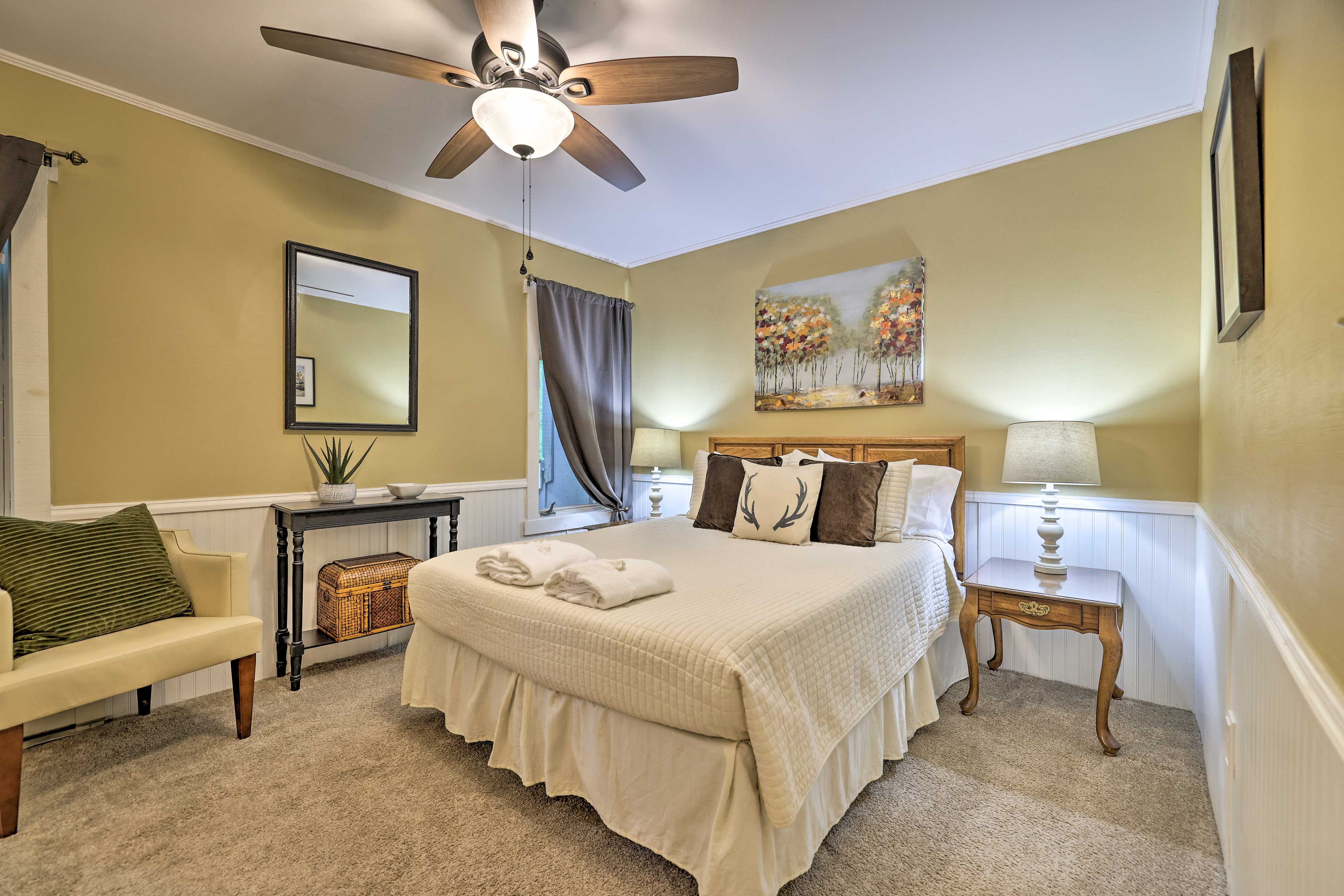 A bedroom with a bed, a nightstand, a chair, a ceiling fan, a painting, and a window with curtains.