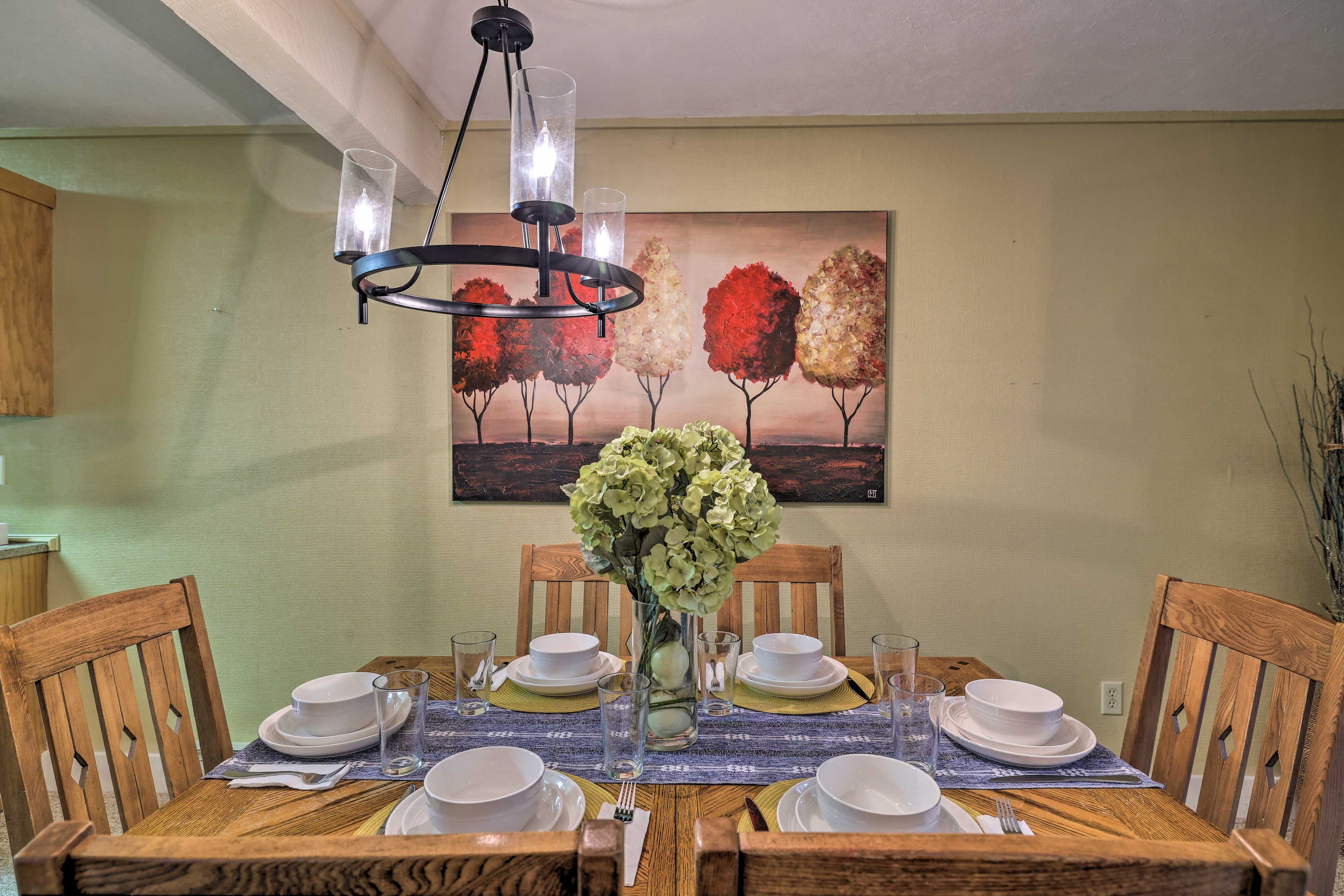 Una mesa de comedor preparada para cuatro personas, con platos, tazones y vasos blancos, un florero con flores y un cuadro de árboles en la pared.