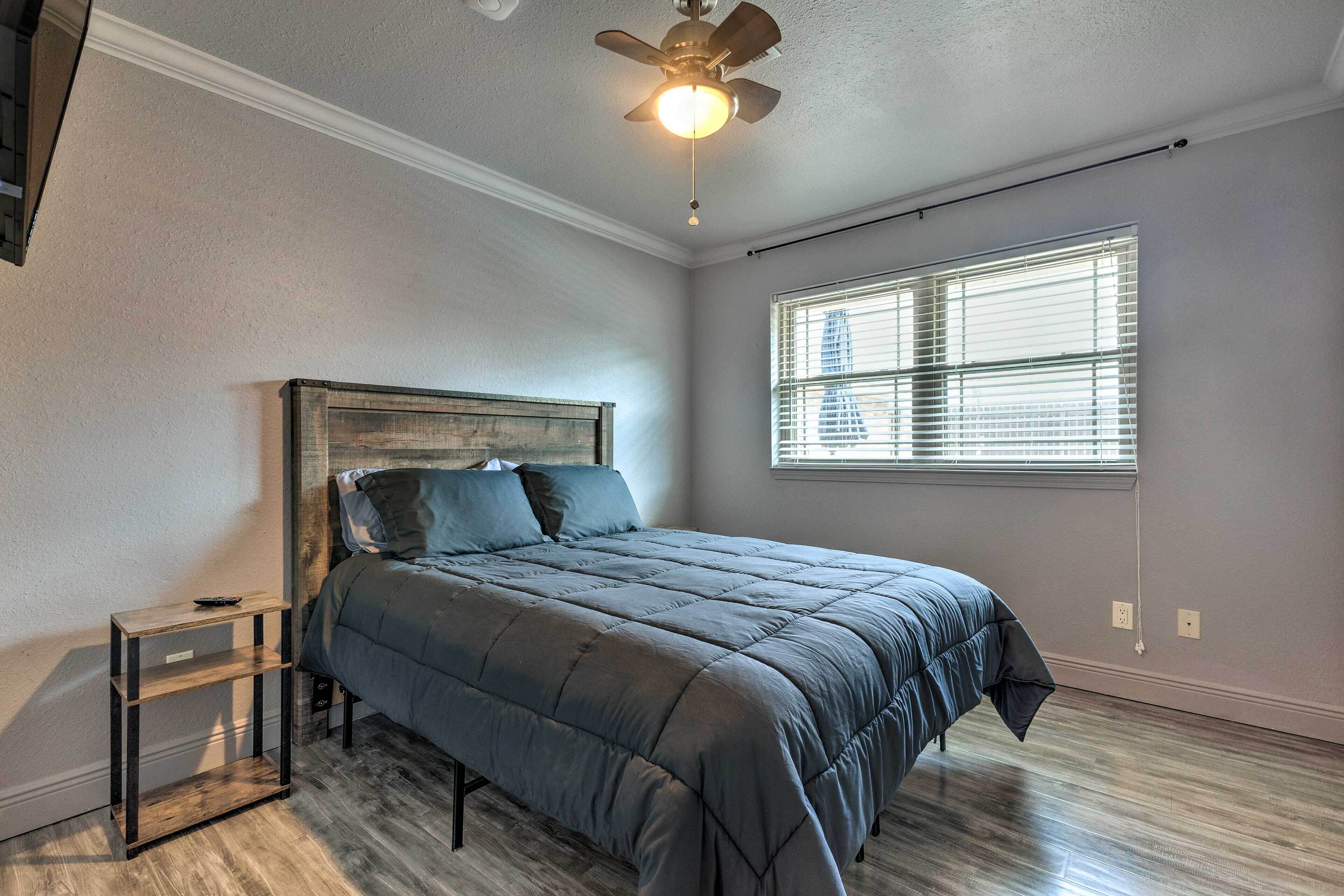 Quarto com cama, mesa de cabeceira, ventilador de teto e janela com persianas.