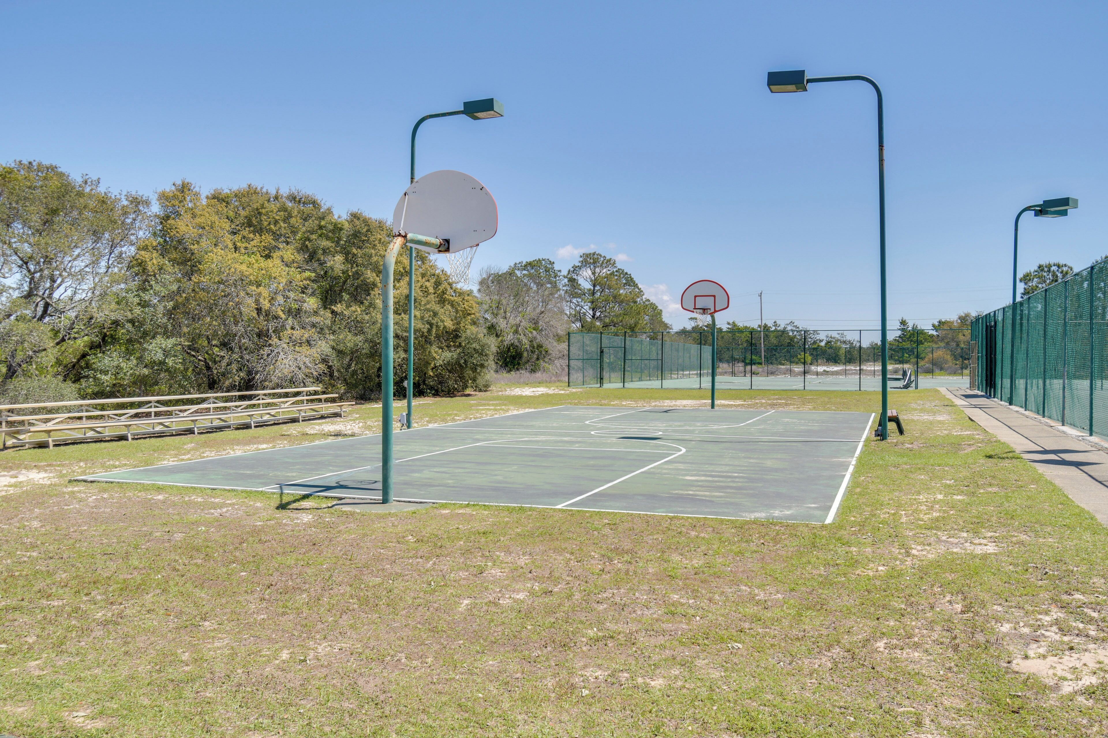 An outdoor basketball court with two hoops, surrounded by a fence and trees.