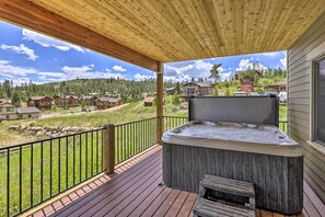 Una terraza de madera con un jacuzzi y un banco, con vistas a una zona residencial con casas y un paisaje montañoso al fondo.