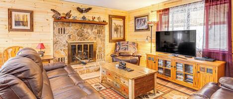 Una sala de estar acogedora con chimenea de piedra, sofás de cuero, una mesa de centro de madera y un televisor sobre un mueble de madera.