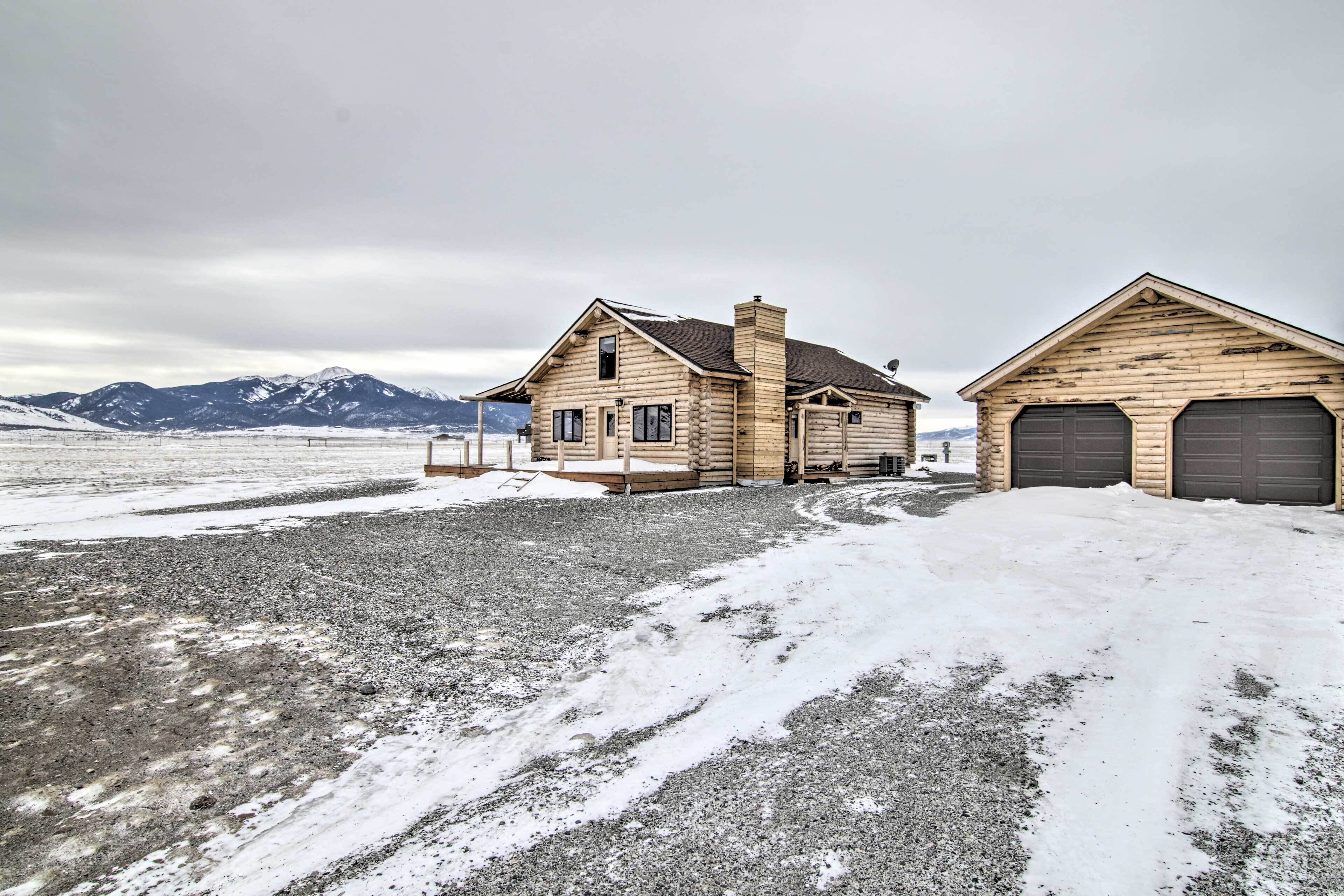 A log cabin with a garage, surrounded by a snowy landscape and mountains in the background.