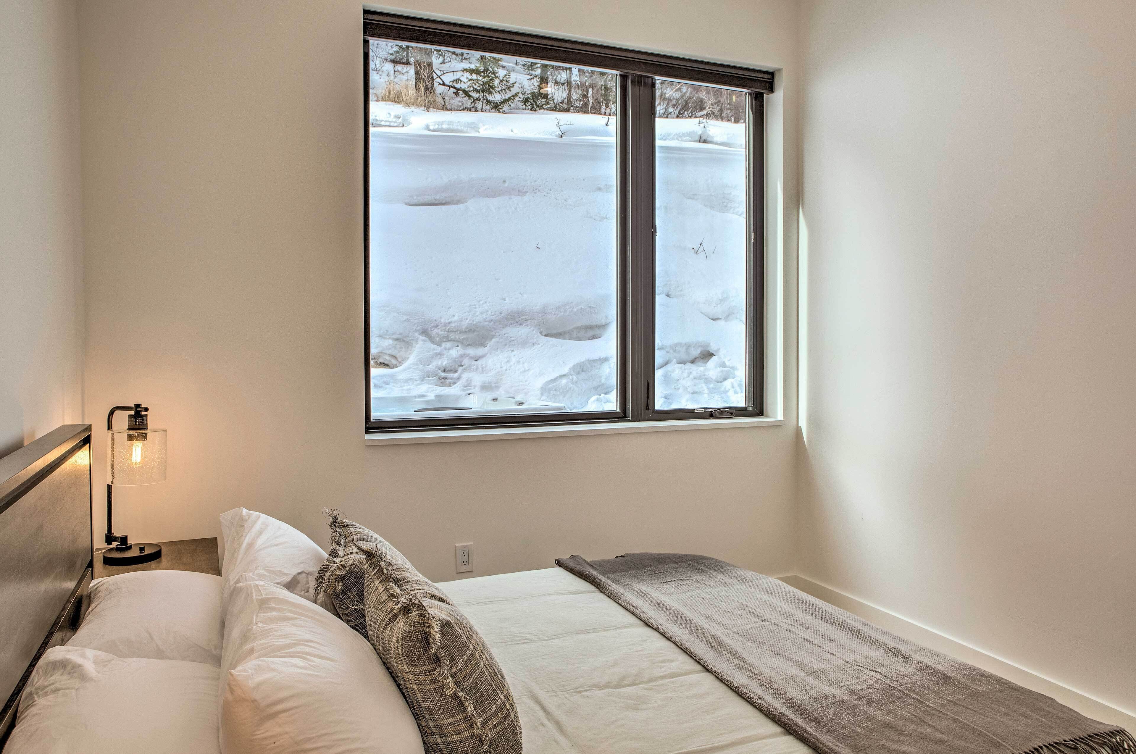 Een slaapkamer met een bed, een raam met uitzicht op een besneeuwde omgeving, een bedlamp en een wandcontactdoos.