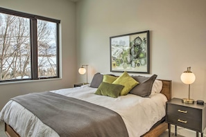 A neatly made bed with a gray and white duvet, two bedside lamps, a framed artwork above the bed, and a window with a view of trees and a snowy landscape.