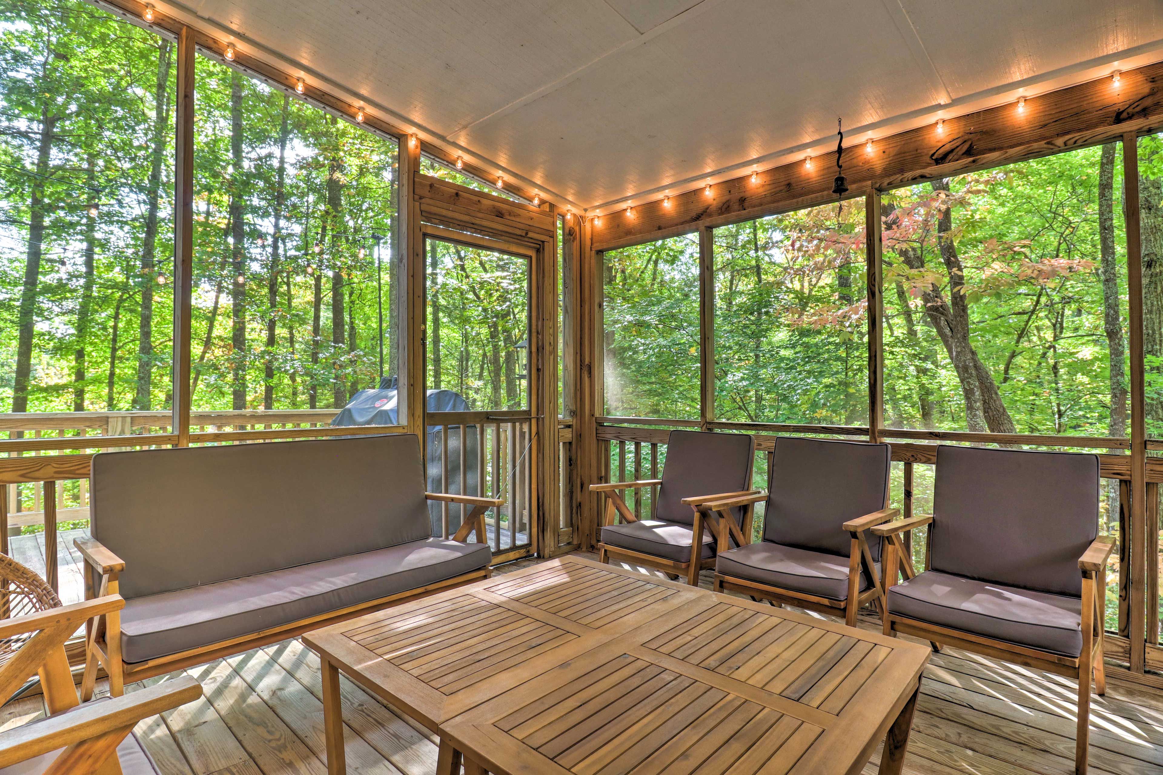A covered outdoor seating area with wooden furniture and large windows overlooking a forest.
