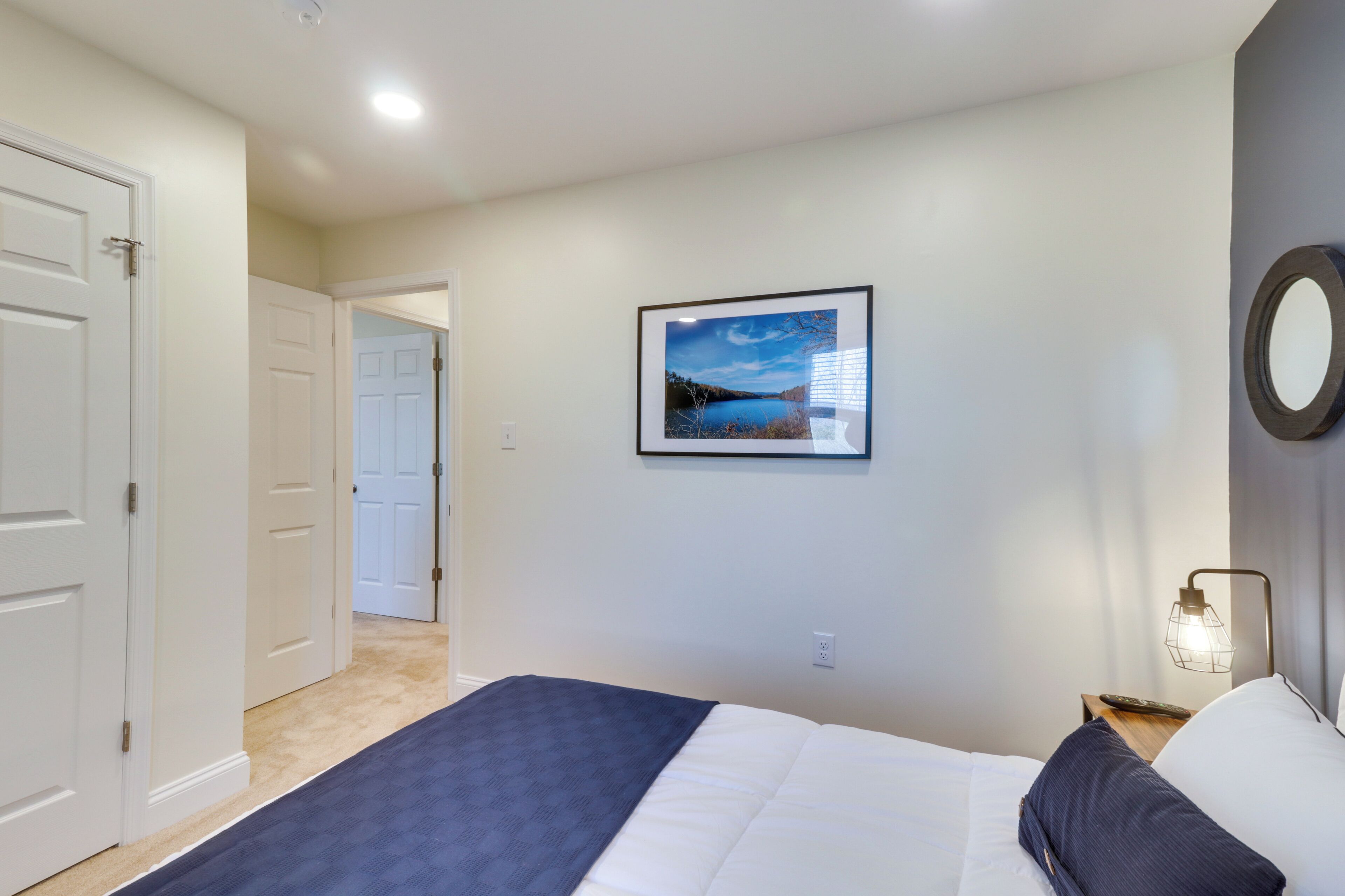 Un dormitorio con cama, mesita de noche, un cuadro enmarcado en la pared y una puerta que da a otra habitación.