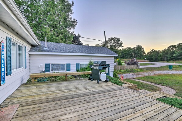 A wooden deck leading to a house with a 'WELCOME' sign, a barbecue grill, and a green trash bin. - East Tawas Cabin w/ Lake Huron Access & Views (East Tawas)
