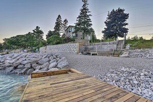 Una terraza de madera que conduce a una costa rocosa con un muro de piedra y una casa al fondo.