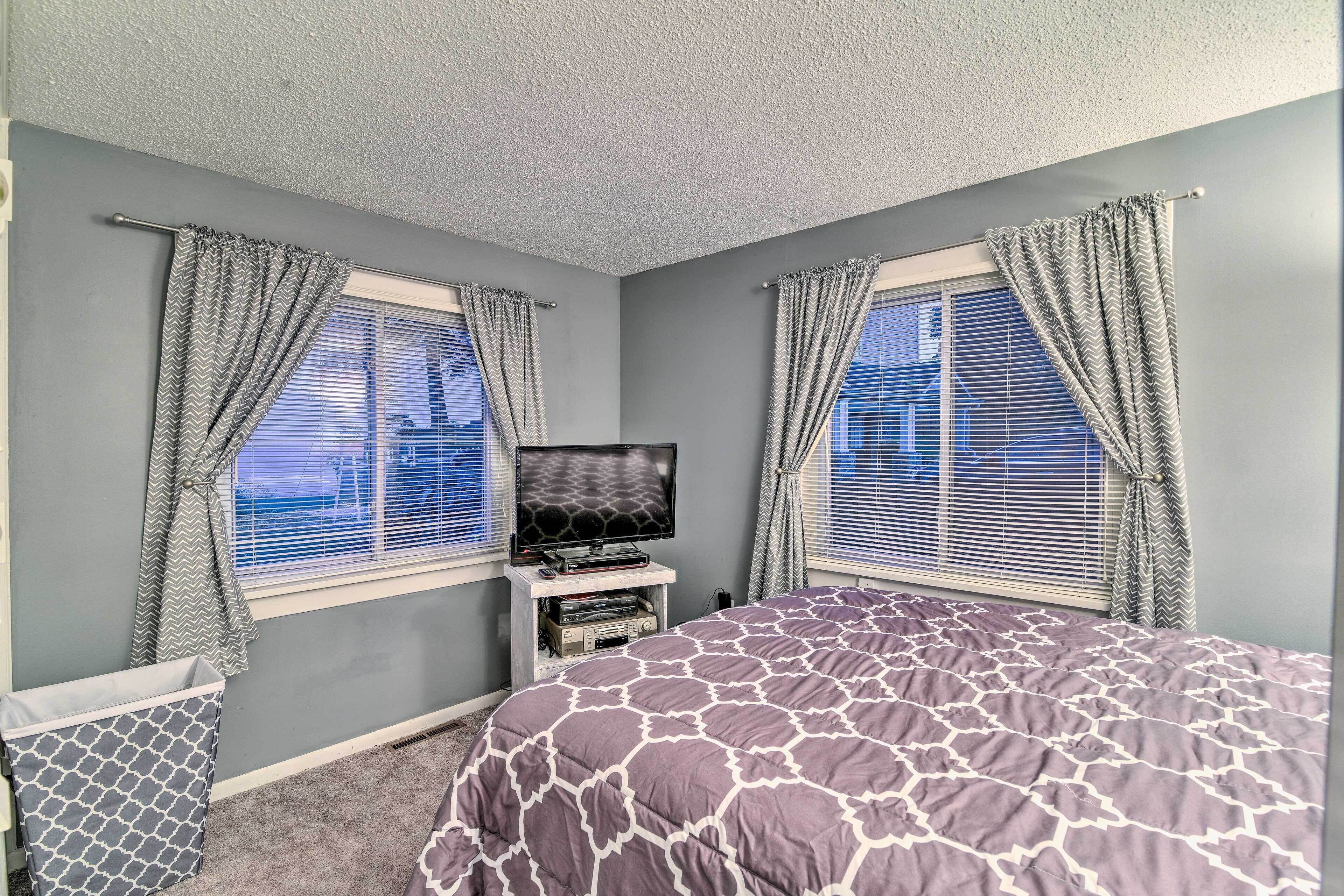 A bedroom with a bed, a television on a stand, and two windows with curtains.