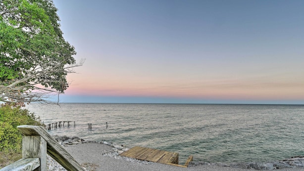 Una terraza de madera que conduce a una playa de guijarros con una plataforma de madera, rodeada de árboles y un mar tranquilo bajo un cielo despejado.