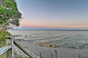 Une terrasse en bois mène à une plage de galets, elle-même surmontée d’une plateforme en bois. L’ensemble est entouré d’arbres et bordé par une mer calme sous un ciel dégagé.