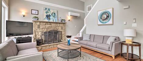 A living room with a stone fireplace, a flat-screen TV, a gray sofa, a round coffee table, and a blue and white rug.