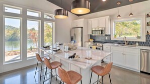 A modern kitchen with a central island, bar stools, and large windows offering views of a lake and trees. - Modern Norfork House w/ Patio on White River! (Norfork)