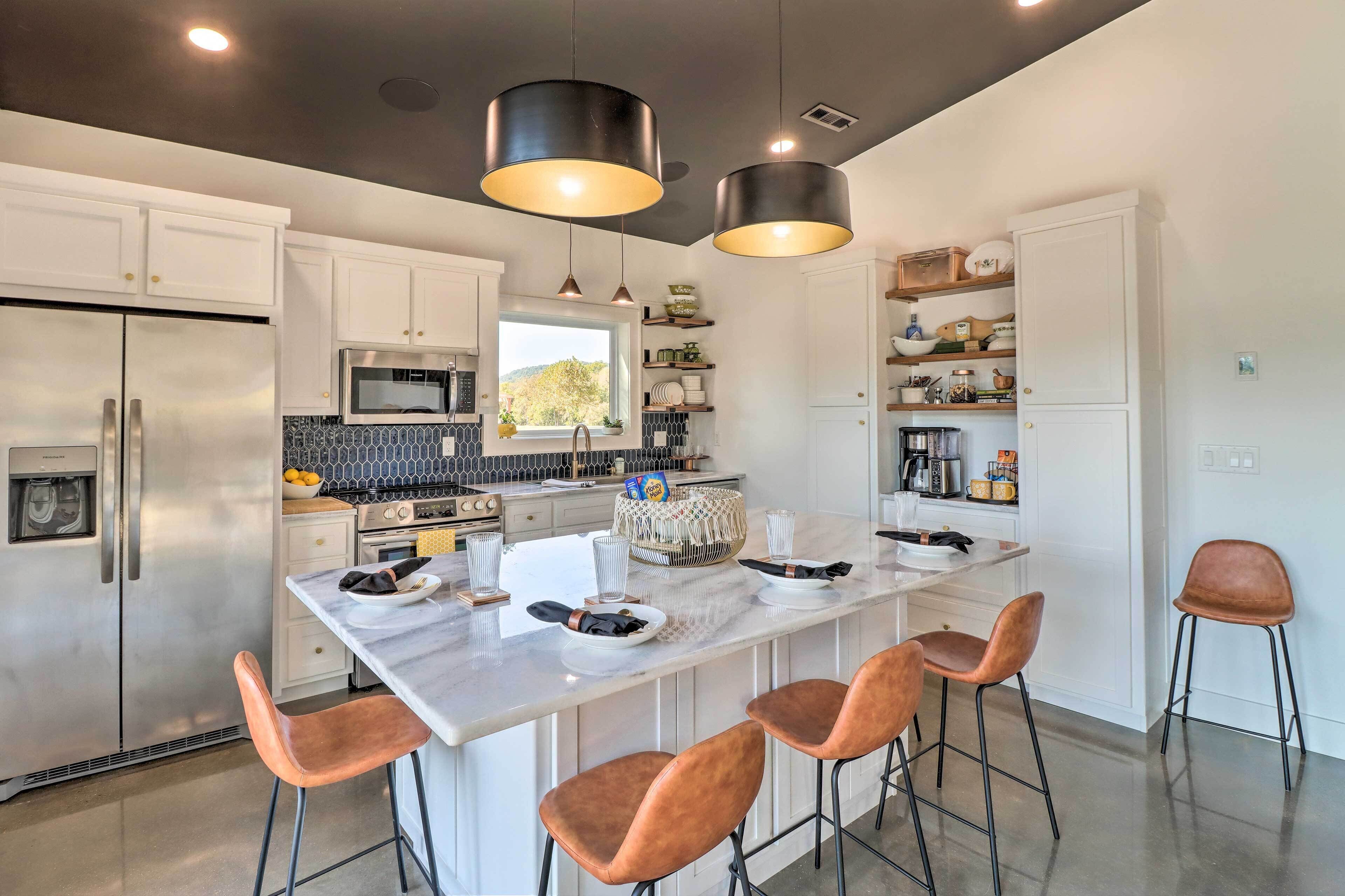 A modern kitchen with white cabinets, stainless steel appliances, a central island with bar stools, pendant lighting, and a view of the outdoors.