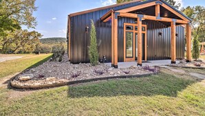 A modern metal building with a wooden porch, surrounded by a landscaped garden with rocks and plants. - Modern Norfork House w/ Patio on White River! (Norfork)