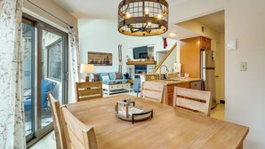 Un coin repas avec une table en bois, des chaises et un lustre. On aperçoit au fond une cuisine équipée d’un réfrigérateur et d’un lave-vaisselle.