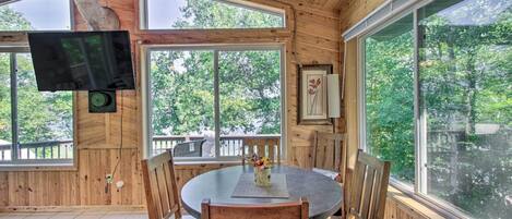 A wooden cabin interior with a round dining table, chairs, a mounted television, and large windows offering a view of the outdoors.