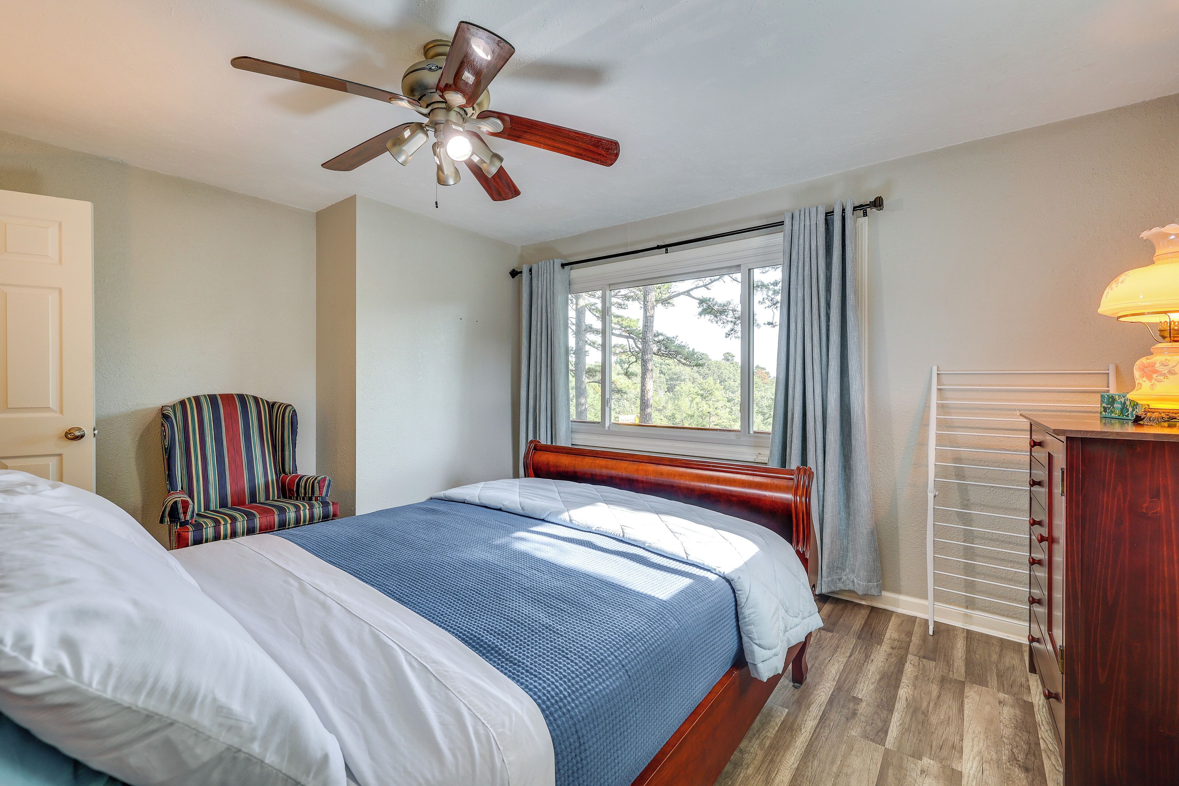 A bedroom with a bed, a ceiling fan, a window with curtains, a chair, and a bedside table with a lamp.