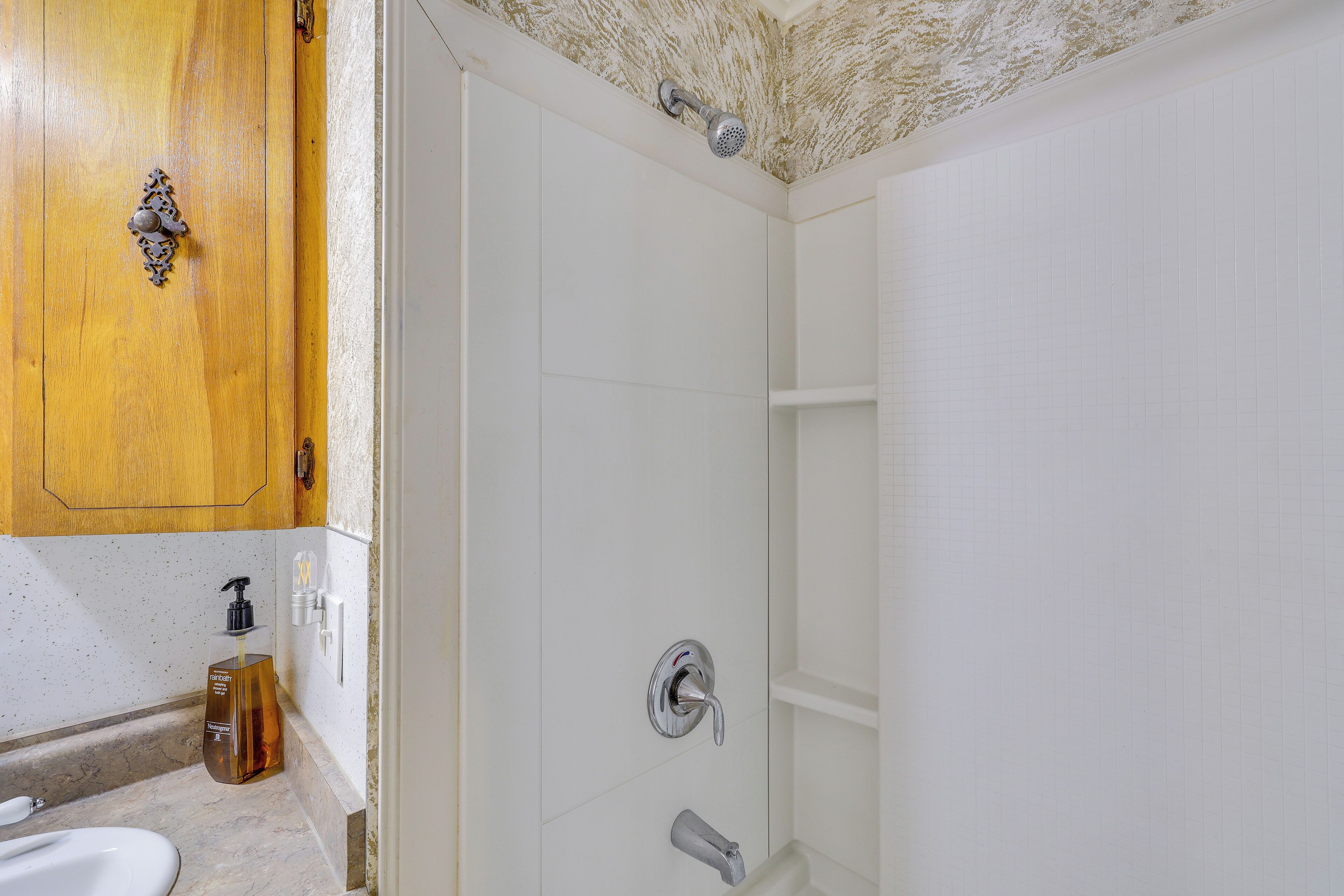 A bathroom with a wooden cabinet, a white tiled wall, a shower with a handheld showerhead, and a soap dispenser on the countertop.