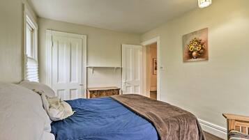 A bedroom with a bed, two doors, a wooden nightstand, a painting, and a chair.
