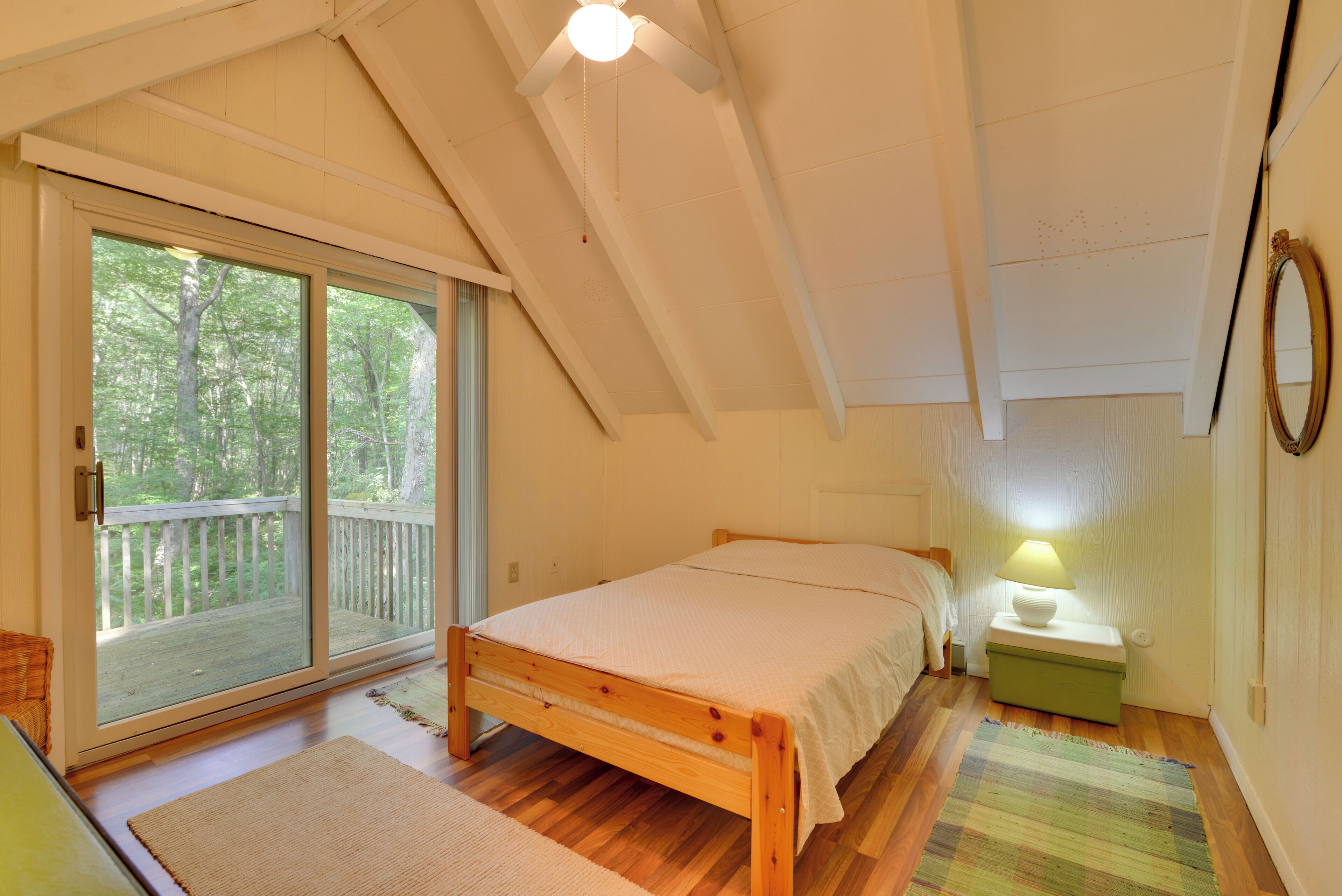 A bedroom with a wooden bed, a nightstand with a lamp, a sliding glass door to a balcony, and a view of trees.