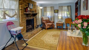 Ett mysigt vardagsrum med en tegelbeklädd öppen spis, ett matbord dukat för två personer och en barnstol vid fönstret.