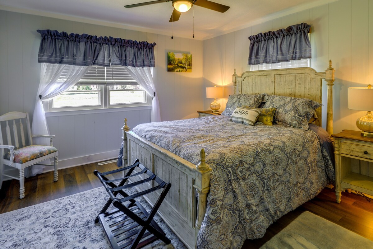 A bedroom with a bed, a chair, a nightstand, a lamp, a ceiling fan, and a window with curtains.