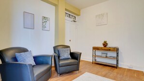 Ein Zimmer mit zwei schwarzen Ledersesseln, einem weißen Teppich, einem Holztischchen mit einer Pflanze und einer weißen Tür.