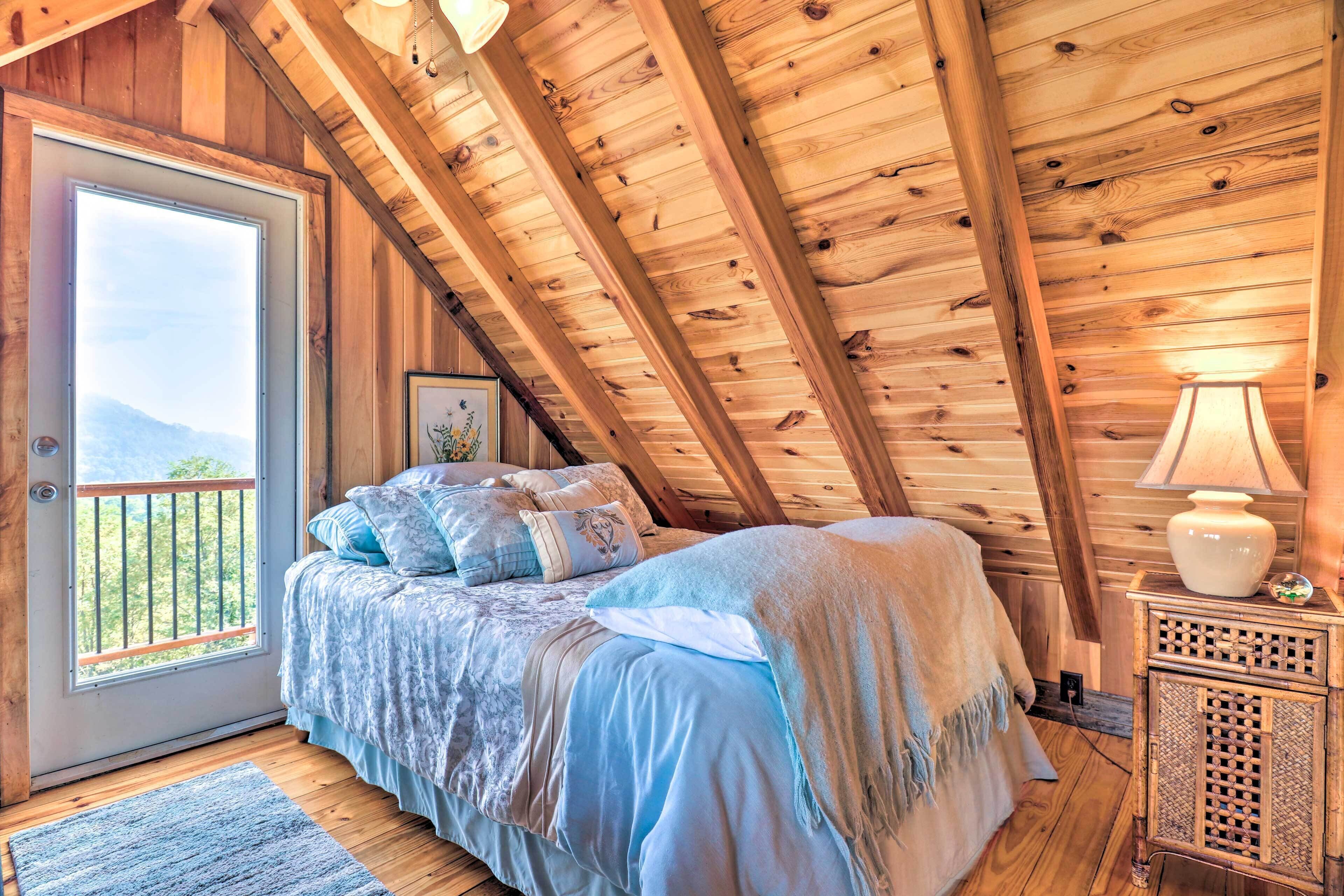 A bedroom with a wooden ceiling, a bed with blue bedding, a nightstand with a lamp, and a window with a view of mountains.