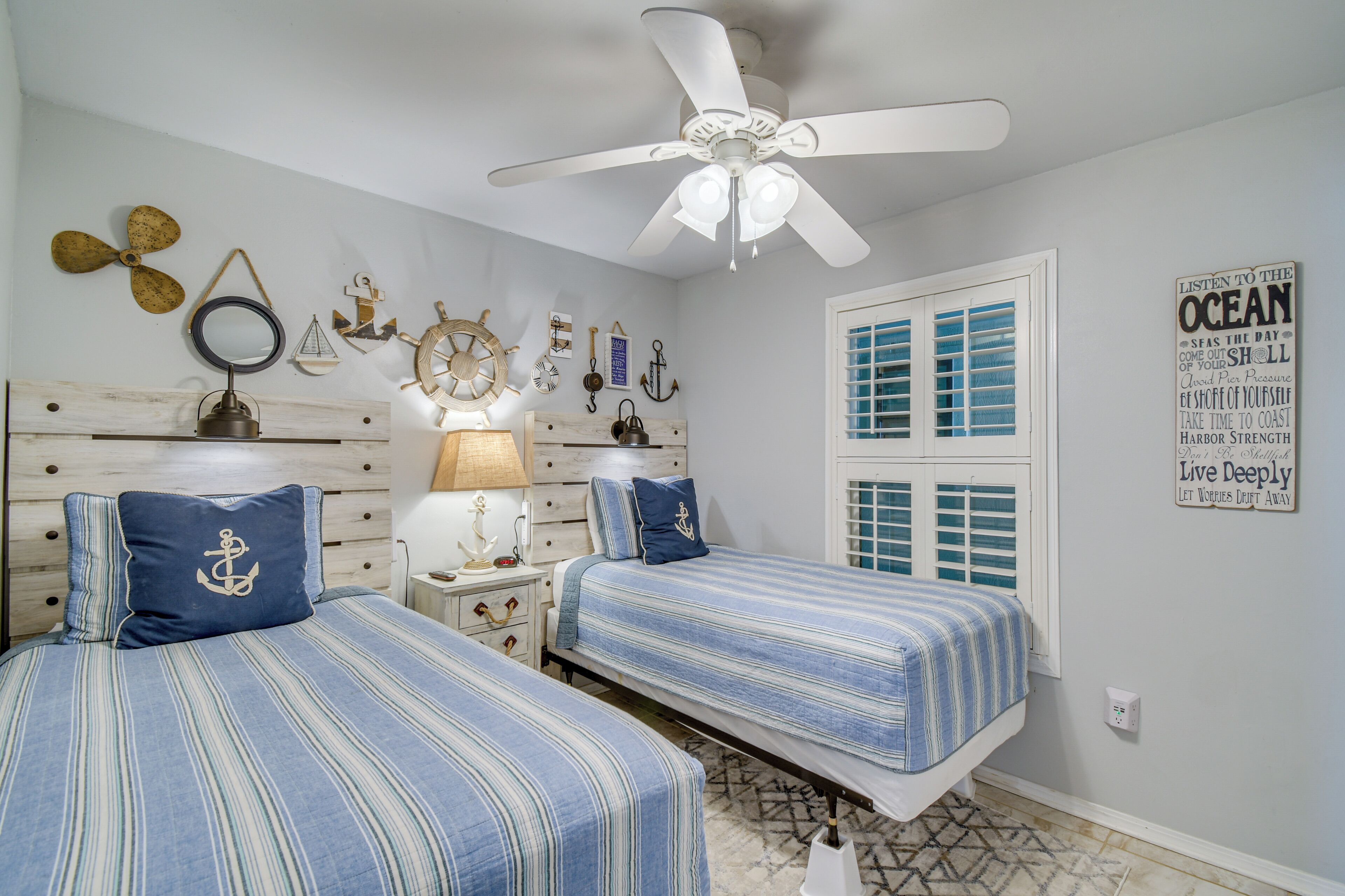 Une chambre avec deux lits, un ventilateur de plafond, une décoration sur le thème de la marine et une fenêtre avec des volets.