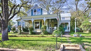 A two-story house with a porch, surrounded by a well-maintained lawn and trees. - Walk to Main St Action! Spacious Historic Retreat (Fredericksburg)