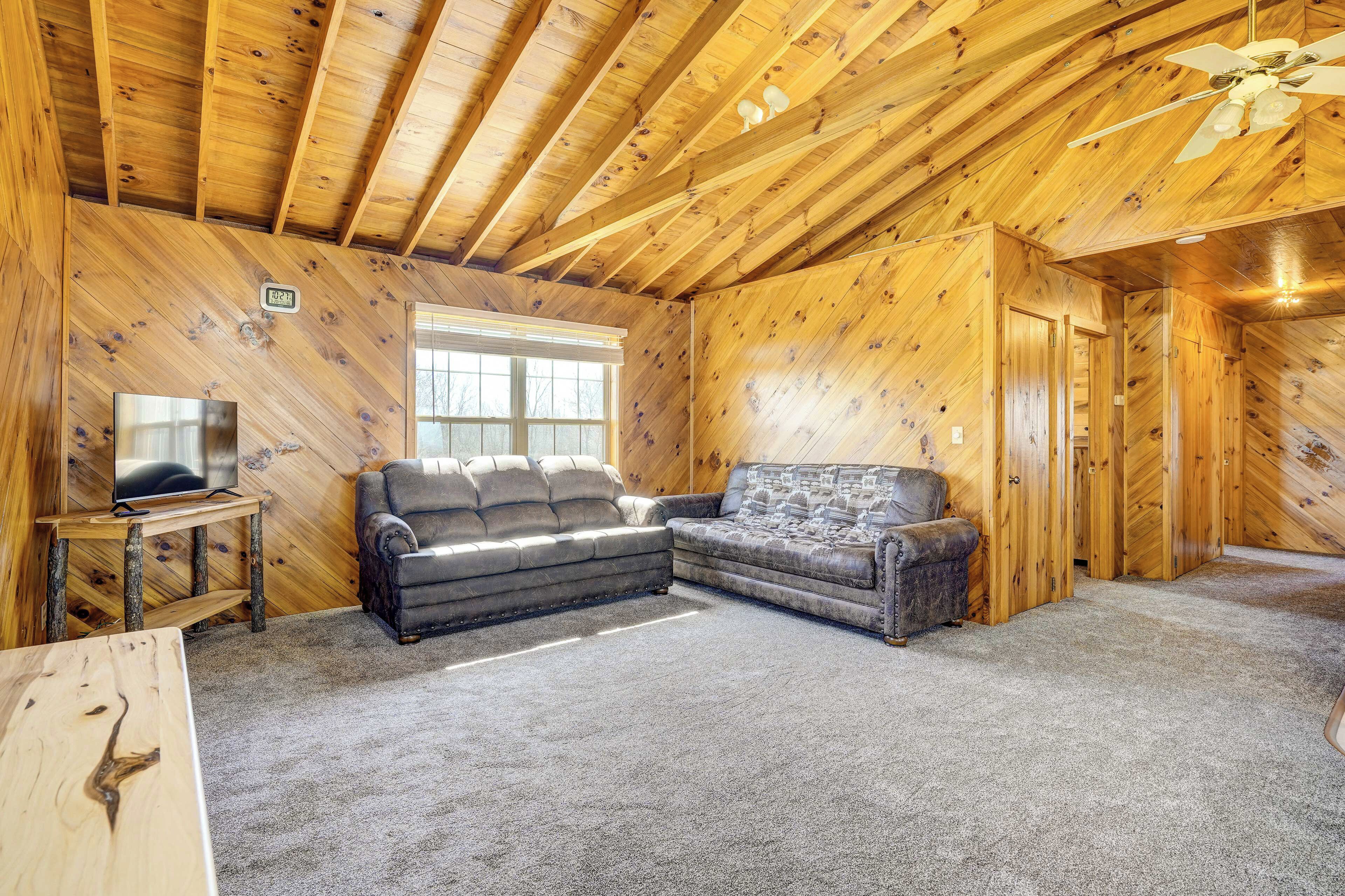 A log cabin interior with a wooden ceiling, a television on a wooden stand, two sofas, and a carpeted floor.